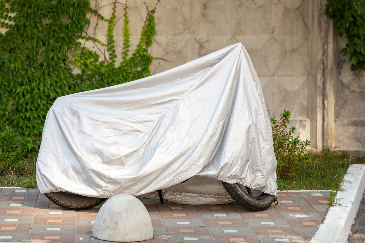 Motorcycle covered with grey protective jacket . Scooter with fabric shield parked at pavement near concrete wall. Waterproof parking outdoors during travel.